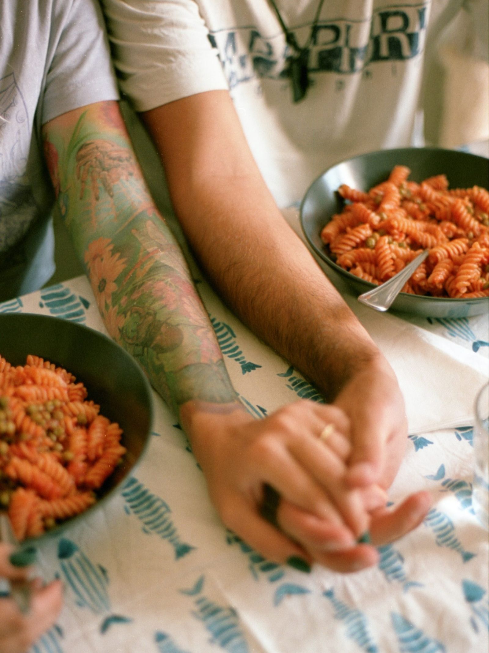 Due persone a tavola, davanti a piatti di pasta, si tengono per mano; uno dei due avambracci è tatuato.