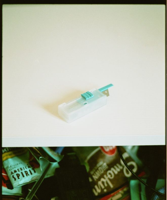 Una scatoletta di lamette su un piano bianco; sotto, un cassetto aperto con tabacco e cartine.