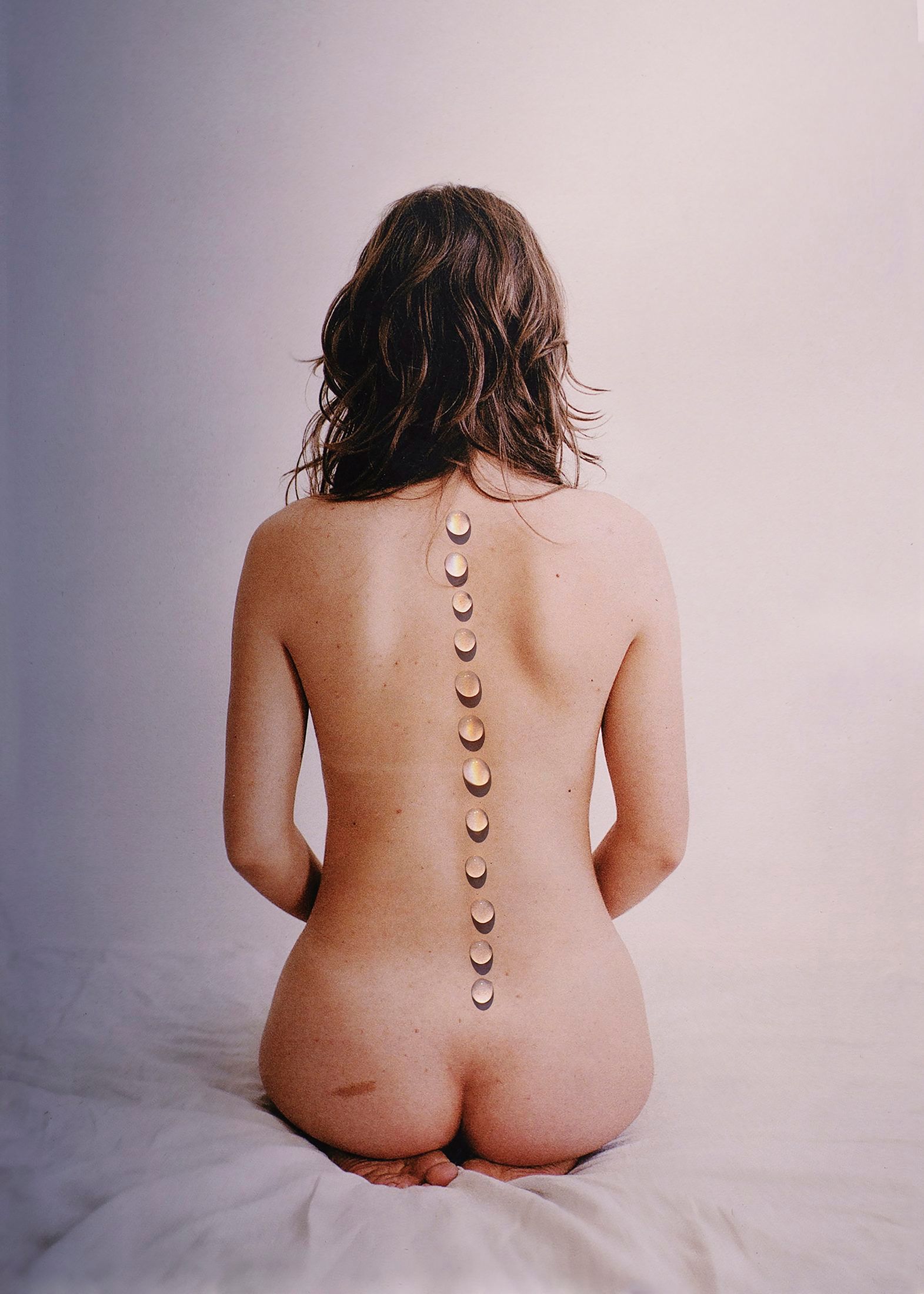 Una donna nuda seduta di schiena; lungo la colonna vertebrale, una fila di gocce d'acqua.
